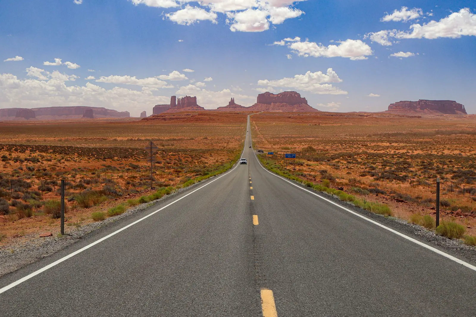 An empty two-lane highway running through red desert rock under a blue sky in Monument Valley, Arizona.